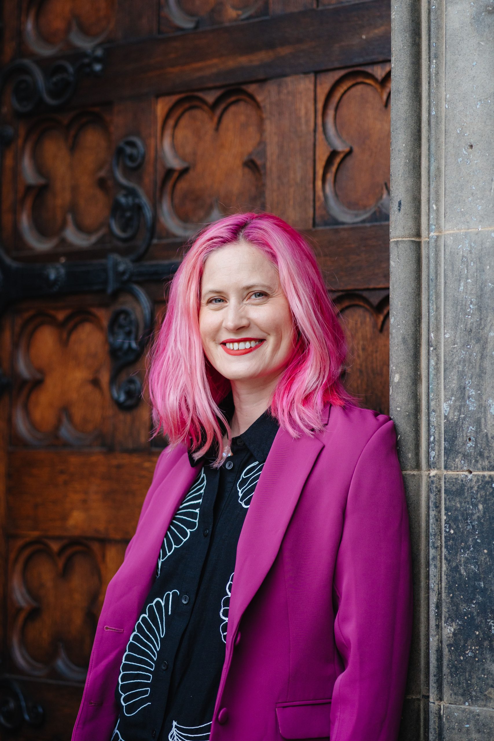 A white woman in her late 30's with bright pink hair and red lipstick. Laura Jayne is smiling. She is standing in front of a cathedral door in Edinburgh.